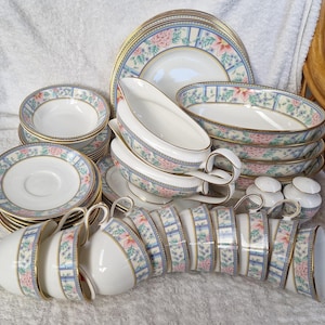 May include: A full porcelain dinner service with a floral design in pink, blue, and gold. The set includes plates, saucers, bowls, cups, serving dishes, and salt and pepper shakers. The edges of the dishes are trimmed with gold.