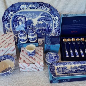 Op de afbeelding: Een collectie Spode Blue Italian serviesgoed, met een grote serveerschaal, zout- en pepervaatjes, een kom, een kleine kop, een dekselschaal, een rechthoekige schaal en lepels. De stukken zijn wit met blauwe landschapsontwerpen.