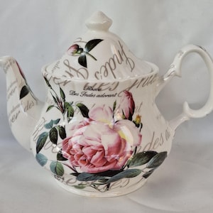 Pode incluir: Bule de cerâmica branca com um grande desenho de rosa rosa e folhas verdes. O bule tem uma alça e um bico curvos, com as palavras "Rose Des Indes adorant" impressas na lateral. A tampa tem um pequeno botão.