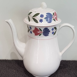 Könnte beinhalten: Weiße Kaffeekanne aus Keramik mit einem Blumendesign in Blau-, Rot- und Grüntönen. Die Kanne hat einen Deckel mit einem Knopf oben.