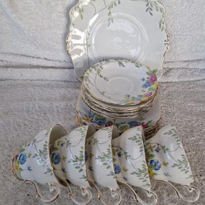 Pode incluir: Um conjunto de chá vintage com chávenas, pires e pratos de porcelana branca. O conjunto é decorado com padrões florais em azul, amarelo e rosa, com acabamentos dourados. Os pratos têm uma borda ondulada. O conjunto inclui chávenas de chá, pires e um prato de servir.