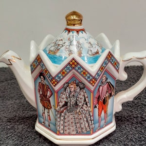 Op de afbeelding: Een zeshoekige keramische theepot met een goudkleurige knop op het deksel. Het lichaam toont kleurrijke illustraties van historische figuren in klederdracht en een bloemenrand. Het deksel toont zeilschepen op een blauwe zee.