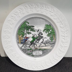 Könnte beinhalten: Ein weißer Keramikteller mit einem erhabenen Rand aus Blättern. Die Mitte des Tellers zeigt eine Schwarzweißillustration einer Szene aus dem Buch "Doctor Syntax" von William Combe. Die Illustration zeigt einen Mann zu Pferd, der von zwei Männern zu Fuß verfolgt wird. Der Text "Doctor Syntax,  Illust. by W. Combe" ist in Schwarz auf dem Teller gedruckt.