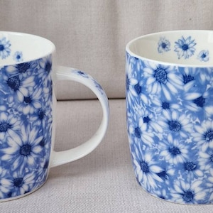 May include: Two white ceramic mugs with a blue floral pattern. The mugs have a white interior and handles. The floral design features white daisy-like flowers on a blue background. The mugs are suitable for hot or cold drinks.
