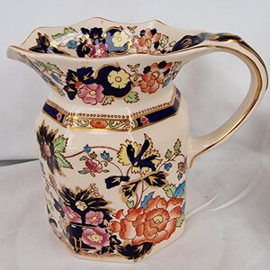 Puede incluir: Jarra de cerámica color crema con diseño floral. La jarra tiene un cuerpo hexagonal y un borde festoneado y acampanado. El patrón floral incluye flores naranjas, rosas y azules con ribetes dorados en el asa y el borde.