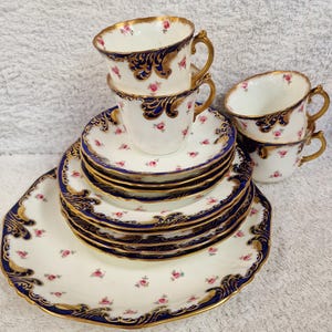 Peut inclure: Service à thé vintage en porcelaine blanche ornée de motifs de roses roses. L'ensemble comprend des tasses, des soucoupes et un plat de service, tous avec des bordures dorées et des accents bleu marine.