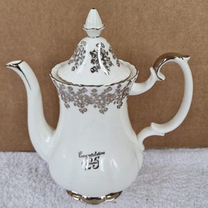 May include: White porcelain teapot with a silver floral design and a silver trim. The teapot has a lid with a silver knob and a silver handle. The teapot has the text "Congratulations 25 years" on the front.