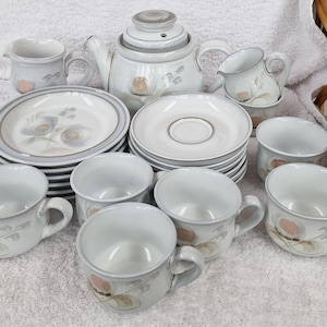 Puede incluir: Un juego de tazas de té, platillos y una tetera de cerámica blanca y gris con un diseño floral. El juego incluye una jarra de crema y un azucarero.