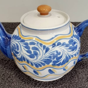Può includere: Una teiera in ceramica bianca con manico e coperchio blu. La teiera ha un design floreale blu e giallo.