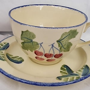 Può includere: Servizio da tè in ceramica color crema. La tazza e il piattino sono decorati con foglie verdi e ciliegie rosse. I bordi sono rifiniti con un bordo blu. La tazza ha un manico curvo.