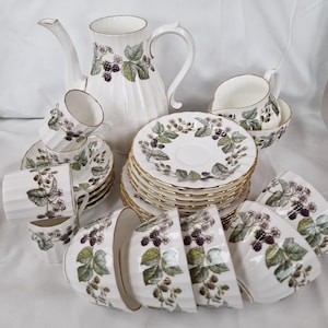 Op de afbeelding: Een vintage witte theeservies met een braambessenontwerp. De set bevat een theepot, kopjes, schotels, kommen en een melkkan. De randen van de kopjes, schotels en kommen zijn goudkleurig afgewerkt. Het braambessenontwerp heeft groene bladeren en paarse bessen.