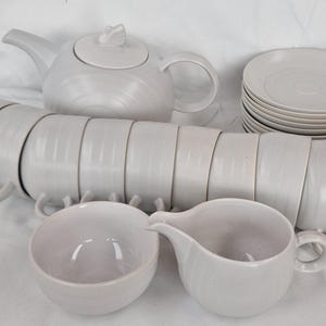 Può includere: Un servizio da tè completo in ceramica bianca, composto da una teiera, tazze, piattini, una lattiera e una zuccheriera. Il set ha un design minimalista con finitura liscia e opaca. Le tazze sono impilate.