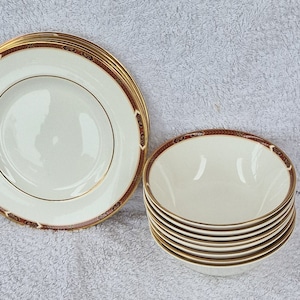 Pode incluir: Um conjunto de tigelas de cerâmica branca e um prato com uma borda dourada e vermelha. As tigelas estão empilhadas e o prato está colocado plano.