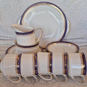 May include: A white porcelain tea set with blue and gold trim. The set includes cups, saucers, a creamer, a sugar bowl, and a large serving plate. The design features a repeating geometric pattern.