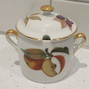 Puede incluir: Azucarero de cerámica blanca con tapa, ribetes y asas doradas. El cuenco está decorado con un colorido diseño de manzanas y hojas. La tapa tiene un pomo dorado y un diseño de moras y hojas.