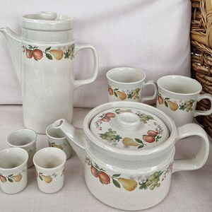 May include: A set of cream-colored ceramic teacups and teapot with a floral pattern featuring pears, apples, and grapes. The set includes a coffee pot, a teapot with a lid, four teacups, and four small cups.