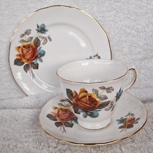 Pode incluir: Um conjunto de chá vintage com uma chávena, um pires e um prato. O conjunto de porcelana branca é adornado com um padrão floral de rosas em tons de laranja, azul e verde, com detalhes dourados. Para servir chá.
