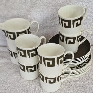 May include: Set of six white ceramic teacups and saucers with a black geometric pattern. The cups and saucers are stacked and arranged on a brown surface.
