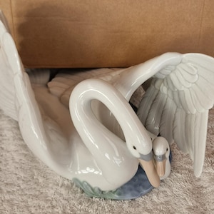 May include: A white ceramic swan figurine with a smaller swan nestled close. The larger swan has outstretched wings, and the smaller swan is tucked in close. The base is blue and green.