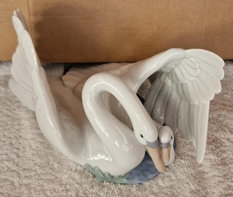 May include: A white ceramic swan figurine with a smaller swan nestled close. The larger swan has outstretched wings, and the smaller swan is tucked in close. The base is blue and green.