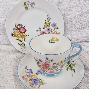 May include: A white teacup, saucer, and plate set with a floral design. The set has a light blue trim and handle. The teacup and saucer have a matching floral pattern with pink, purple, yellow, and blue flowers.