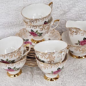Könnte beinhalten: Vintage Teeservice mit weißen Porzellantassen, Untertassen und Sahnekännchen. Das Set ist mit Goldverzierungen, rosa Rosen und goldenen Akzenten verziert. Die Tassen haben einen Fuß und verzierte Griffe. Ein klassisches Teeservice.