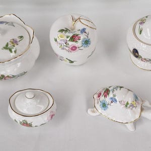 May include: A trio of cream-glazed ceramic mugs with a speckled finish. Each mug is adorned with a hand-painted design: a blue and green leaf, a yellow flower, and a red berry. The mugs have a simple, rounded form.