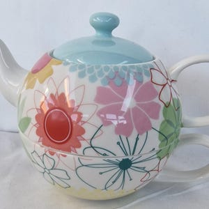 May include: A ceramic tea-for-one set featuring a white teapot with a light blue lid and a matching teacup. The exterior is decorated with a colorful floral pattern in shades of pink, red, yellow, green, and teal, with both pieces stacked together.