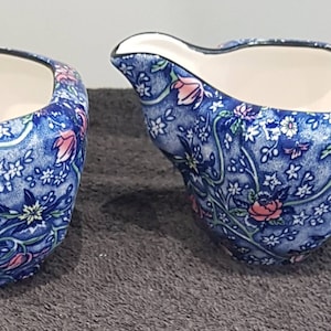 May include: A ceramic creamer and sugar bowl set featuring a vibrant blue base with a dense, intricate pattern of pink flowers, white blossoms, and green vines. Both pieces have a smooth white interior and a distinct, irregular, organic shape.