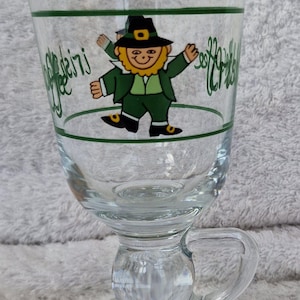 Op de afbeelding: Heldere Irish coffee mok van glas met een handvat en een ronde voet. De mok heeft een groene lijn en een cartoon kabouter illustratie. De woorden "Seoiri" en "Happy" staan in groene letters op het glas.