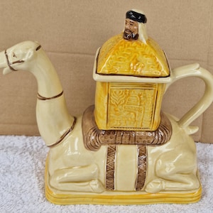 May include: A ceramic teapot shaped like a camel carrying a small house on its back. The house has a brown roof and yellow walls with intricate designs. The camel is cream colored with brown accents.