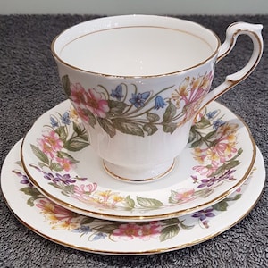 以下が含まれることがあります： 白地のティーカップ、ソーサー、サイドプレートのセット。ピンク、黄色、青の花柄のデザインで、金色の縁取りが施されています。ティーカップはカーブしたハンドルが付いています。