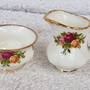 Puede incluir: Un juego de azucarero y crema de porcelana blanca. El juego presenta un diseño floral con rosas rojas, rosas y amarillas, acentuadas con hojas verdes. El borde dorado bordea las piezas, agregando un toque de elegancia.
