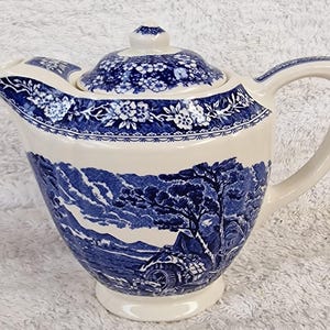 May include: Cream ceramic teapot with a blue and white landscape design. The teapot has a lid with a floral pattern and a curved handle. The body of the teapot depicts a rural scene with trees and a house.