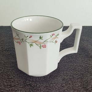 Puede incluir: Taza de té de cerámica blanca con un borde verde y un patrón floral con flores rosas y hojas verdes. La taza tiene una forma cuadrada con bordes redondeados y un asa.