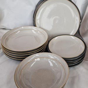 May include: A collection of ceramic plates and bowls. The plates and bowls have a speckled, off-white surface with a dark gray rim. The bowls and plates are stacked in several piles, showcasing a complete dinnerware set.