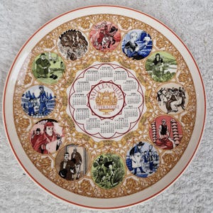 May include: A decorative plate featuring a calendar for the year 1999, with illustrations of literary figures and scenes. The plate has a white background with a red rim and gold accents. The calendar includes names like Oscar Wilde and Charles Dickens.