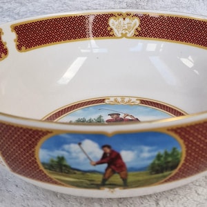 May include: A white ceramic bowl with a burgundy and gold rim. The bowl features golf-themed illustrations and a repeating burgundy pattern. The interior has a glossy finish. The bowl's design includes images of golfers in action.