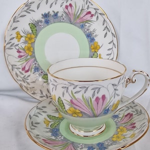 May include: A vintage teacup, saucer, and side plate set. The white porcelain features a floral design with pink, yellow, and blue flowers, accented by gold trim. The saucer and side plate have a scalloped edge.