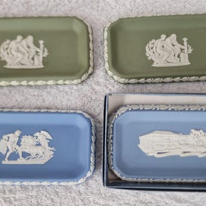 Peut inclure: Quatre petits plats rectangulaires en céramique avec des motifs en relief blancs sur fond vert et bleu. Les motifs comprennent un couple tenant un bâton, un char tiré par un cheval et un bâtiment avec un toit.