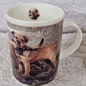 Puede incluir: Taza de cerámica blanca con una ilustración detallada de un perro sosteniendo un palo. La taza tiene un asa y una imagen más pequeña del perro dentro del borde. El perro es marrón y negro, sobre un fondo natural.
