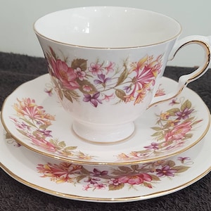 May include: A white teacup, saucer, and side plate set. The set features a floral design in pink, purple, and yellow, with gold trim. The teacup sits on a small footed base, and the handle is also trimmed in gold. The set is designed for tea service.