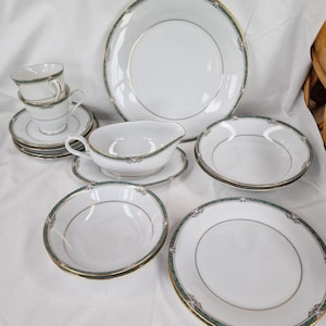 Könnte beinhalten: Ein weißes Porzellan-Tafelservice mit Goldrand und einer grün-goldenen Zierbordüre. Das Set umfasst Teller, Schalen, Untertassen, Tassen und eine Sauciere. Das Geschirr ist auf einer weißen Oberfläche angeordnet.