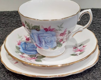 May include: A white teacup, saucer, and side plate set with a blue rose floral design and gold trim. The teacup has a curved handle and sits atop the saucer, with a matching side plate below. The set is designed for tea parties.