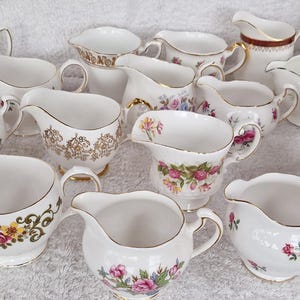 Può includere: Una collezione di 15 brocche per panna in ceramica bianca con dettagli floreali e dorati. Le brocche sono disposte a semicerchio su una superficie bianca.