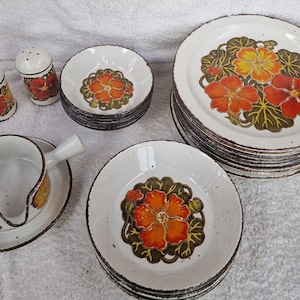 Op de afbeelding: Een vintage servies met witte borden, kommen en een juskom, versierd met oranje, gele en groene bloemmotieven. De set bevat zout- en peperstrooiers. De randen hebben een bruine, getextureerde rand.