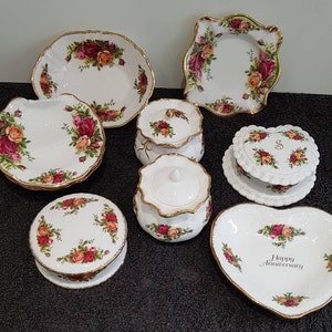 Puede incluir: Un juego de platos de porcelana blanca con ribetes dorados y diseños florales. El juego incluye un plato en forma de corazón con las palabras "Feliz Aniversario" en él, un plato redondo con un diseño floral, un plato cuadrado con un diseño floral y dos tarros con tapa con diseños florales.