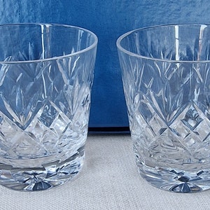 Puede incluir: Dos vasos transparentes de cristal tallado con un patrón de diamantes. Los vasos tienen una boca ancha y una base gruesa. El fondo es de color azul liso. Estos vasos son adecuados para servir bebidas.