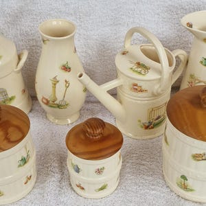 May include: A set of seven ceramic kitchen canisters with wooden lids. The canisters feature a white background with a floral design and a variety of garden-themed illustrations. The canisters are in various shapes and sizes, including a pitcher, a watering can, and a milk jug.