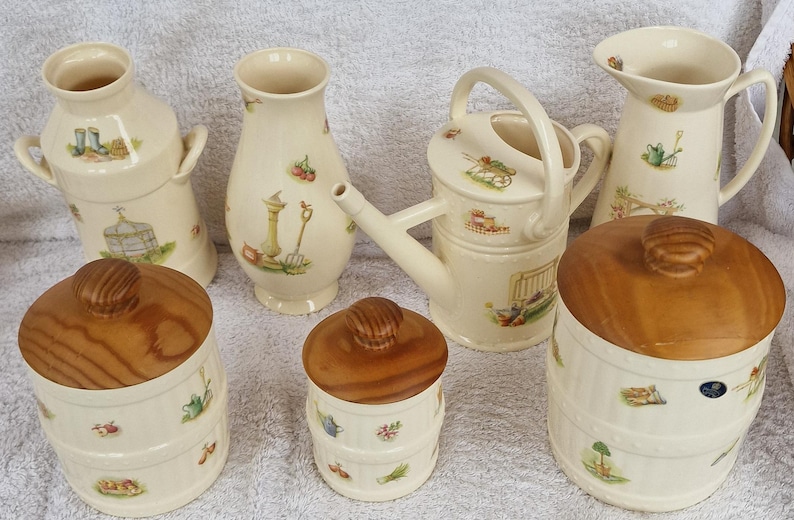 May include: A set of seven ceramic kitchen canisters with wooden lids. The canisters feature a white background with a floral design and a variety of garden-themed illustrations. The canisters are in various shapes and sizes, including a pitcher, a watering can, and a milk jug.